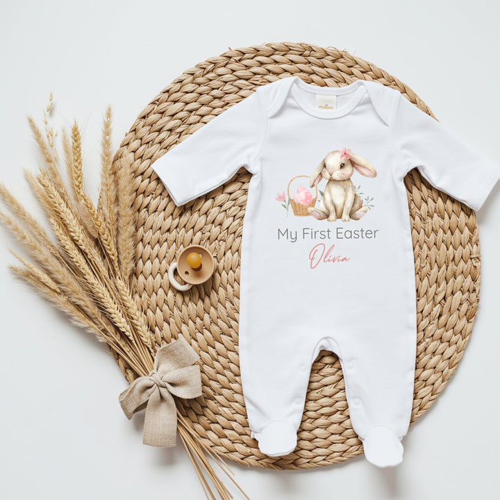 Personalised white Easter baby grow/sleepsuit that says 'My First Easter Olivia'. This design features a brown bunny wearing a pink bow sitting next to a basket with pink eggs in it