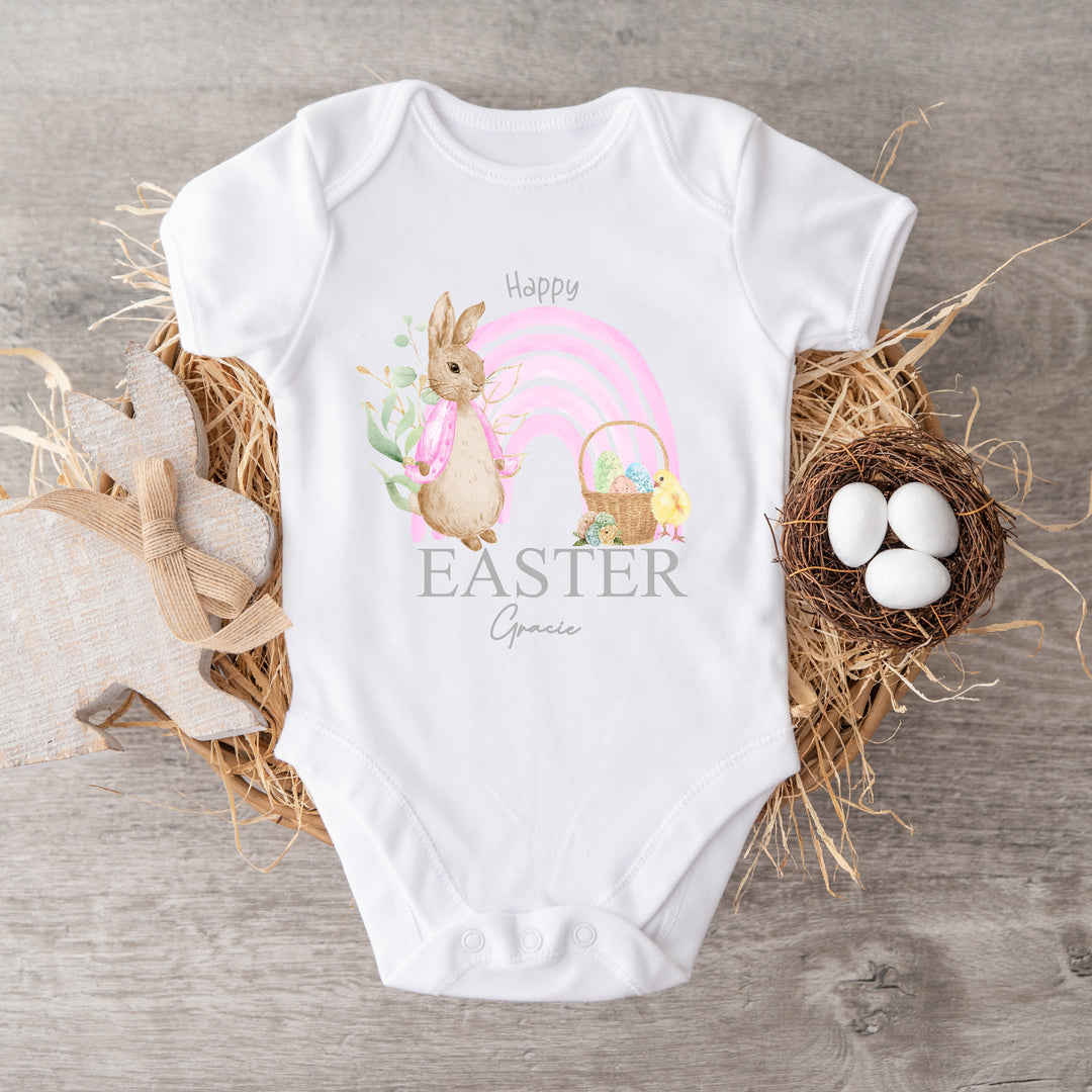 Personalised white Easter baby vest that says 'Happy Easter Gracie'. This design features a pink rainbow with a rabbit wearing a pink jacket one side and an Easter basket and a chick the other