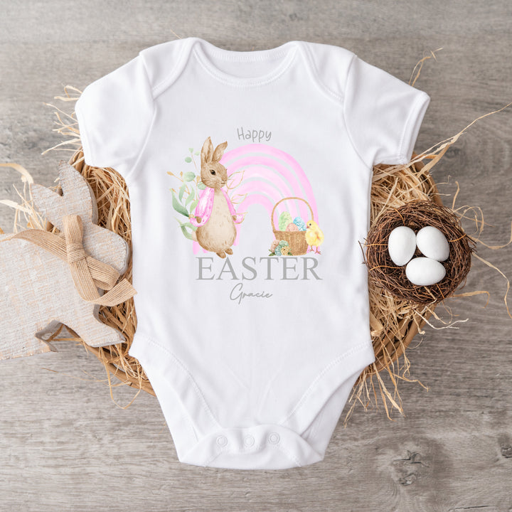 Personalised white Easter baby vest that says 'Happy Easter Gracie'. This design features a pink rainbow with a rabbit wearing a pink jacket one side and an Easter basket and a chick the other