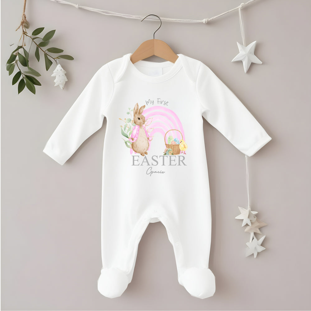 Personalised white Easter baby grow/sleepsuit that says 'My First Easter Gracie'. This design features a pink rainbow with a rabbit wearing a pink jacket one side and an Easter basket and a chick the other