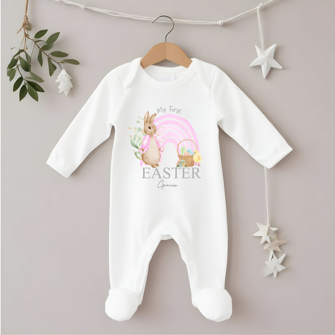 Personalised white Easter baby grow/sleepsuit that says 'My First Easter Gracie'. This design features a pink rainbow with a rabbit wearing a pink jacket one side and an Easter basket and a chick the other