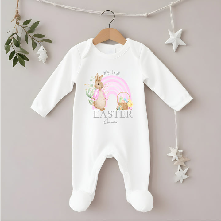 Personalised white Easter baby grow/sleepsuit that says 'My First Easter Gracie'. This design features a pink rainbow with a rabbit wearing a pink jacket one side and an Easter basket and a chick the other