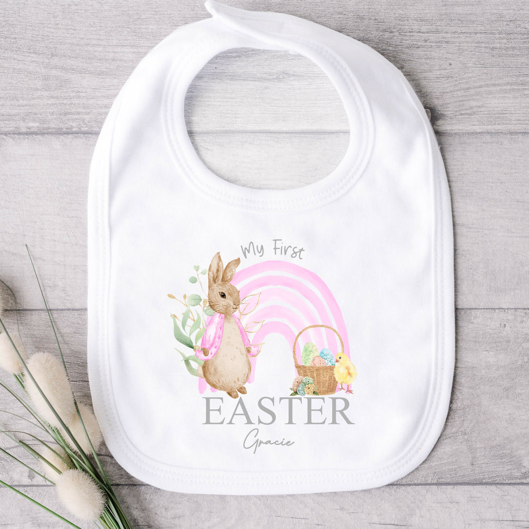 Personalised white Easter baby bib that says 'Happy Easter Gracie'. This design features a pink rainbow with a rabbit wearing a pink jacket one side and an Easter basket and a chick the other