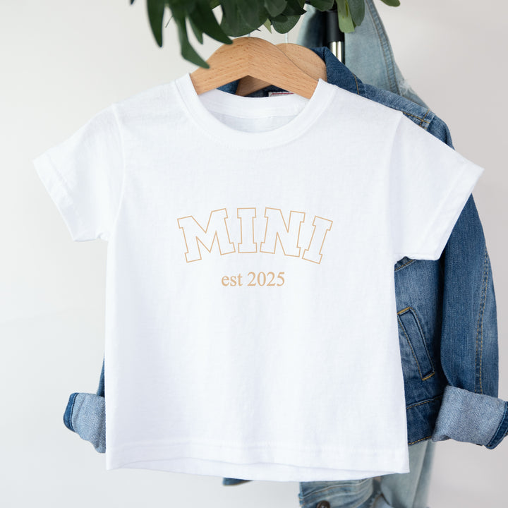White t-shirt with 'MINI est 2025' text on a hanger with denim jacket.