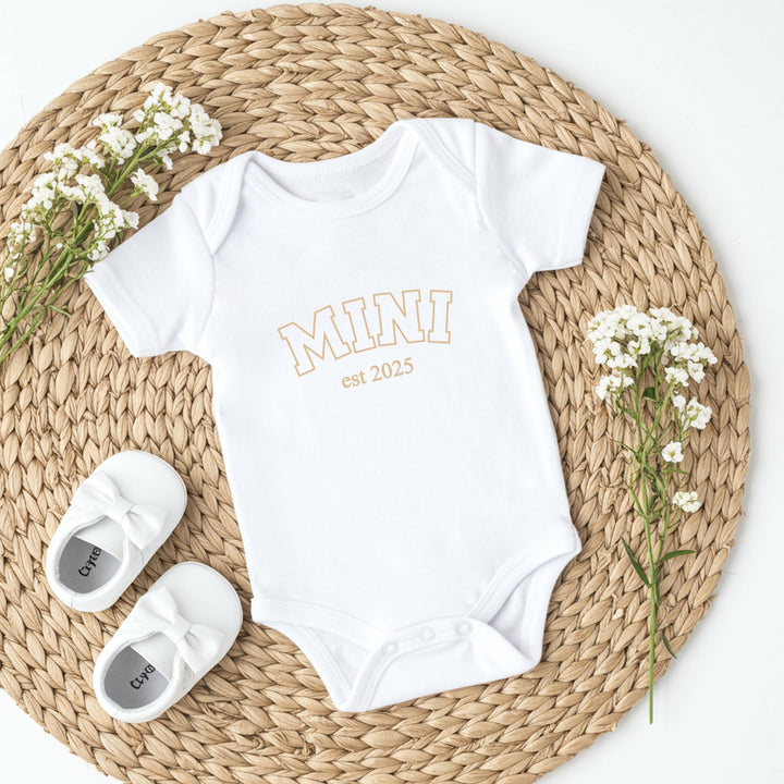 White baby onesie with 'MINI est 2023' text and white baby shoes on a woven mat with flowers.