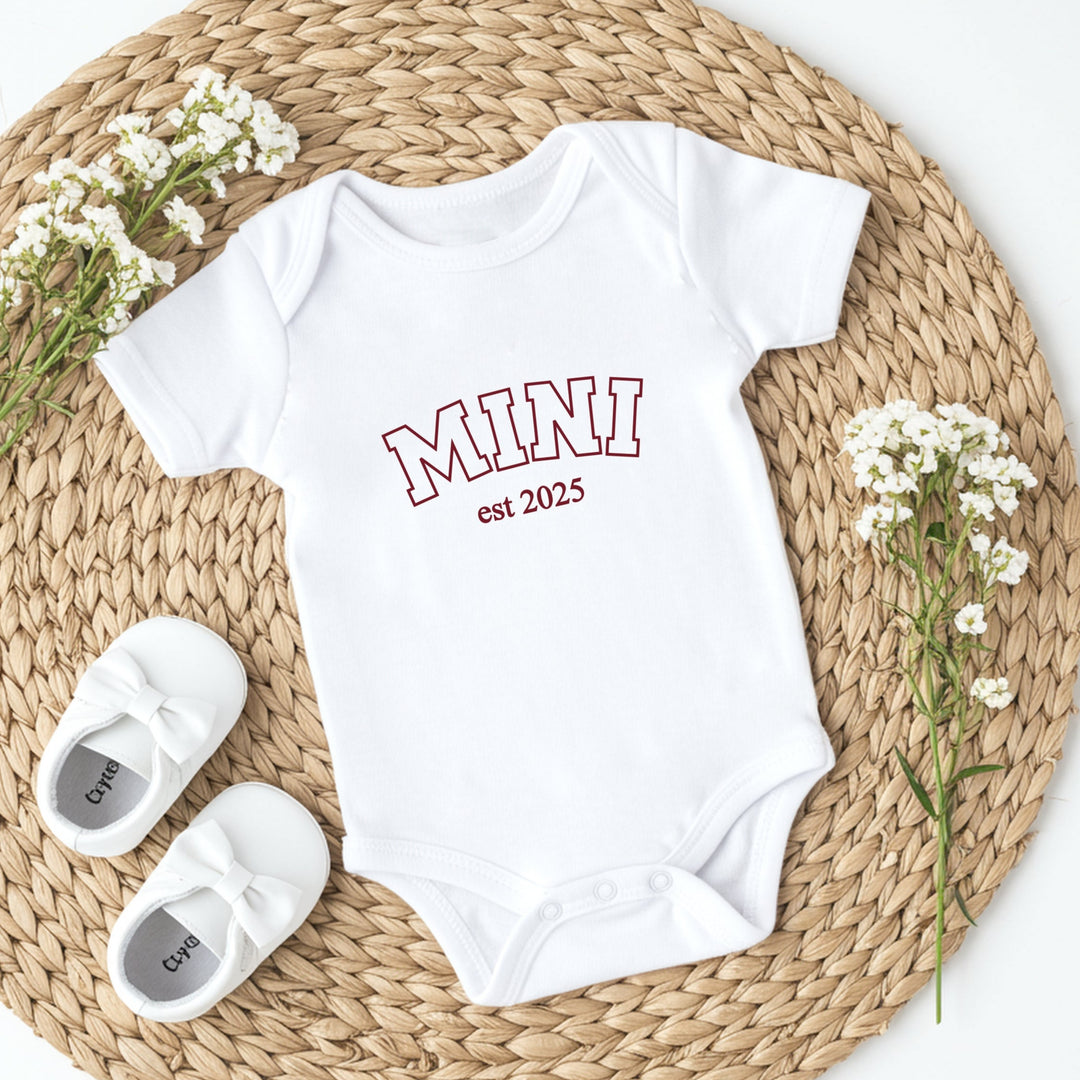 White baby onesie with 'MINI est 2023' text, white baby shoes, and baby's breath flowers on a woven mat.