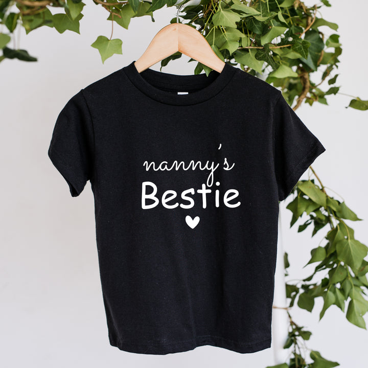 Black t-shirt with 'nanny's Bestie' text on a hanger against a white background with green leaves.