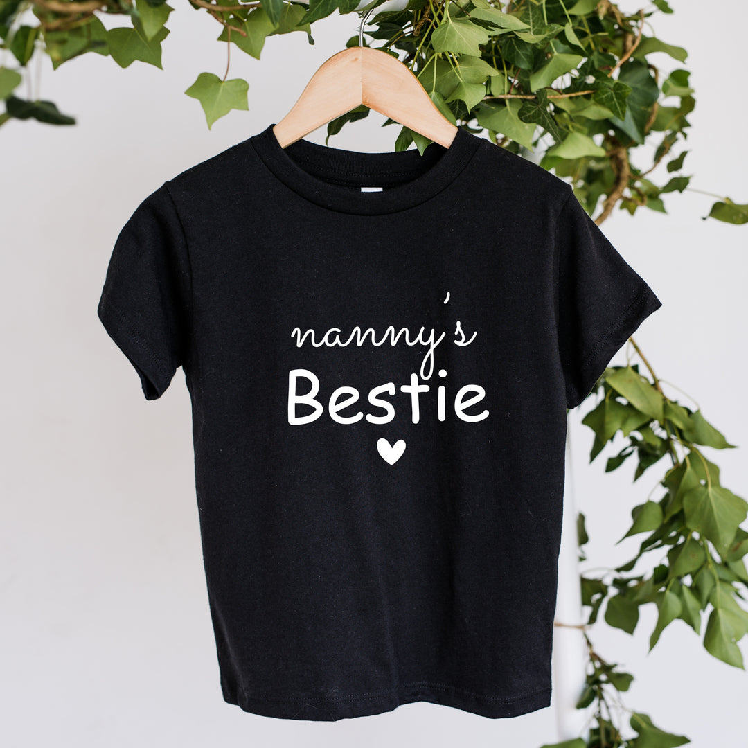 Black t-shirt with 'nanny's Bestie' text on a hanger against a white background with green leaves.