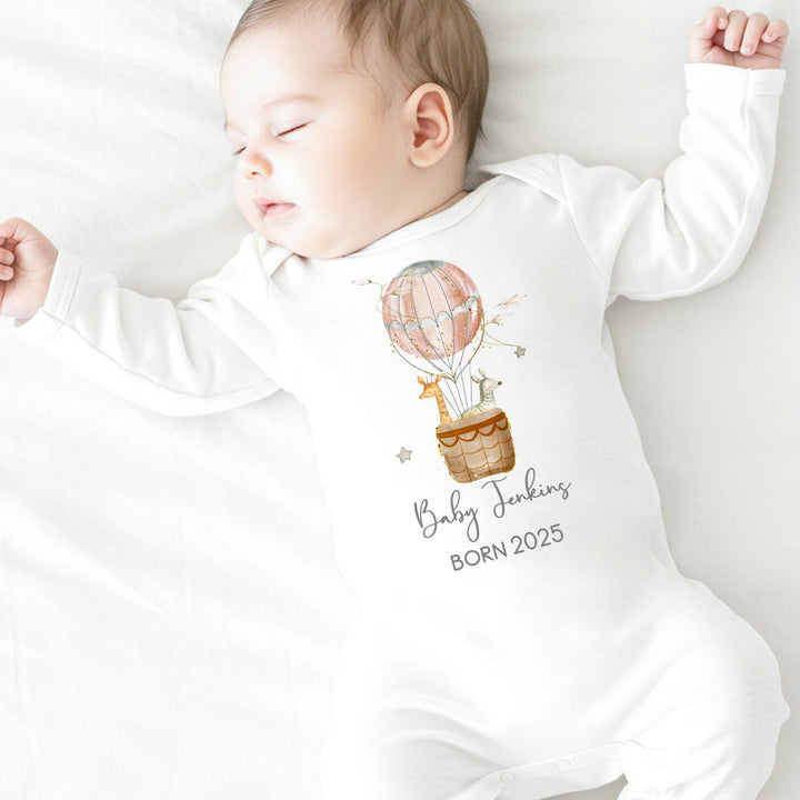 a personalised white baby grow that says 'Baby Jenkins Born 2025'. This design features a pink hot air balloon with a giraffe and zebra in it