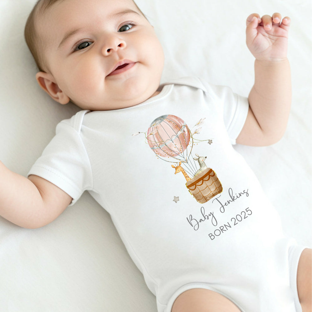 a personalised white baby vest that says 'Baby Jenkins Born 2025'. This design features a pink hot air balloon with a giraffe and zebra in it