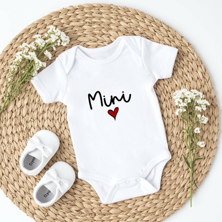 White baby onesie with 'Mimi' and a heart, white shoes, and flowers on a woven mat.