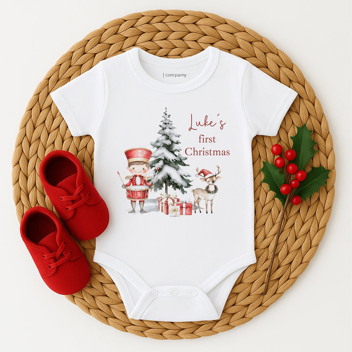 Baby onesie with Christmas design and text, red shoes, and holly on a woven mat.