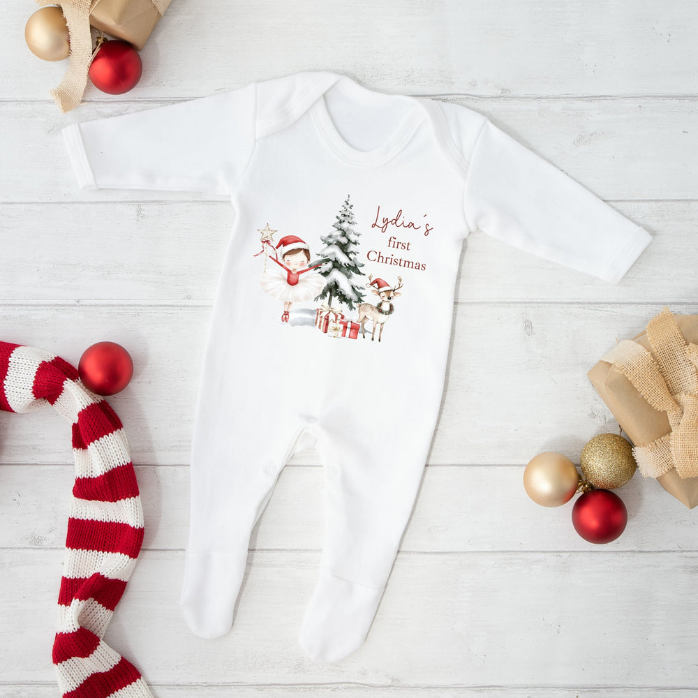 White baby onesie with Christmas design and text, surrounded by festive decorations on a light wooden floor.