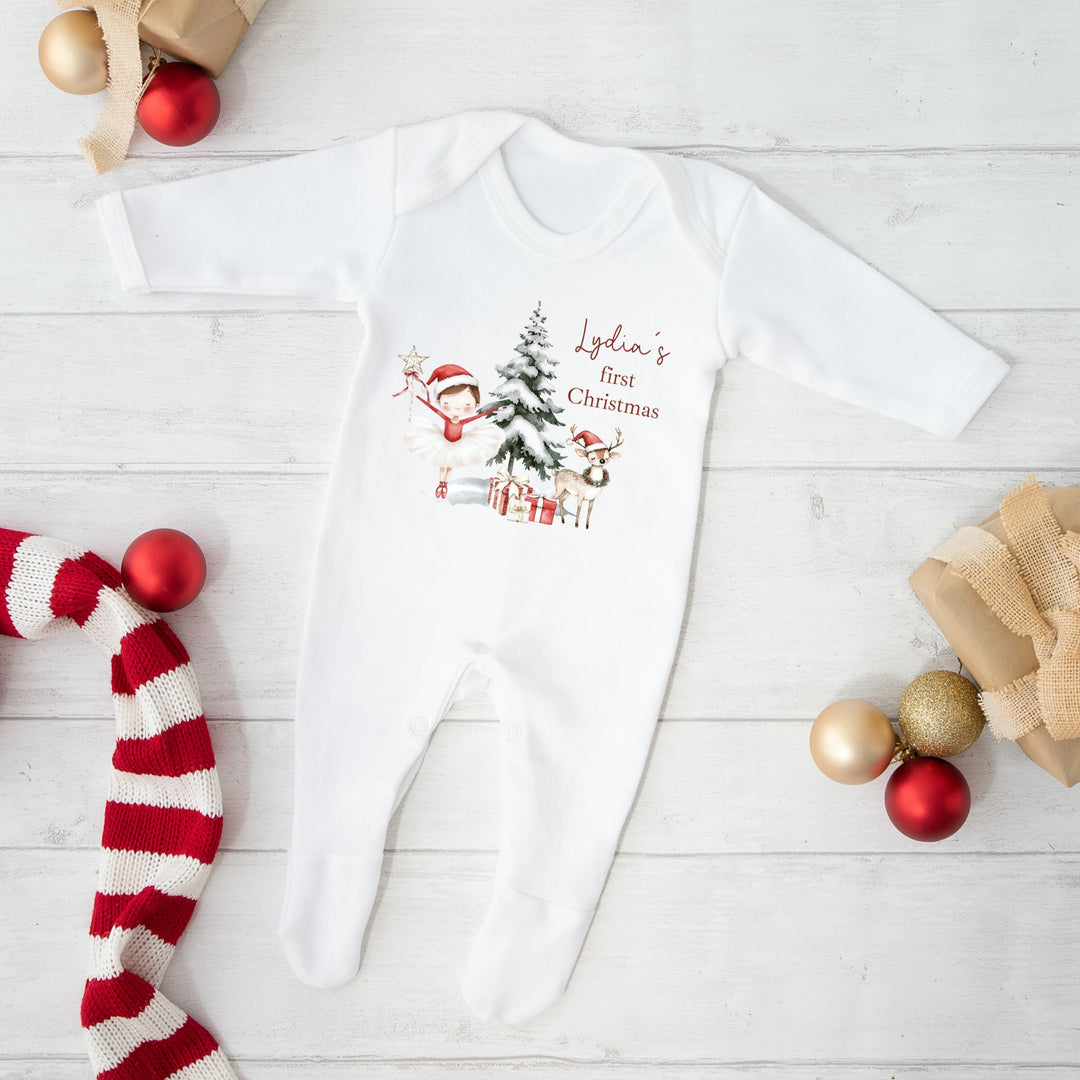 White baby onesie with Christmas design and text, surrounded by festive decorations on a light wooden floor.