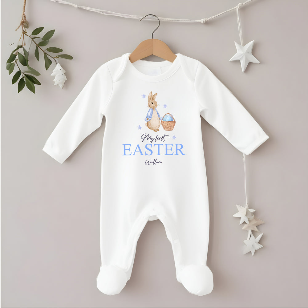 Personalised white Easter baby grow/sleepsuit that says 'My First Easter Wallace'. This design features a rabbit with a blue jacket holding an Easter basket with blue stars around it
