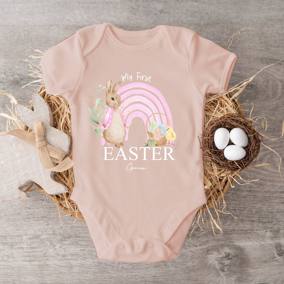 Personalised pink Easter baby vest  that says 'My First Easter Gracie'. This design features a pink rainbow and a rabbit wearing a pink jacket