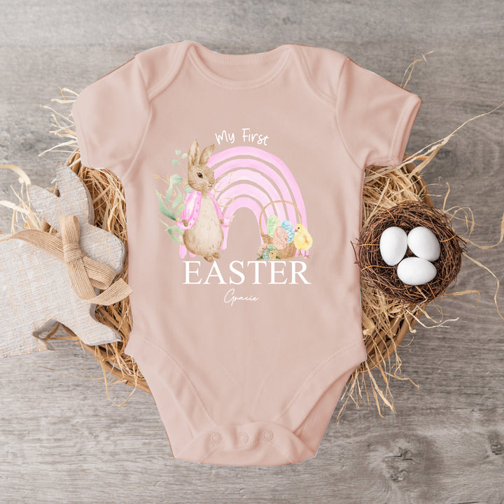 Personalised pink Easter baby vest  that says 'My First Easter Gracie'. This design features a pink rainbow and a rabbit wearing a pink jacket