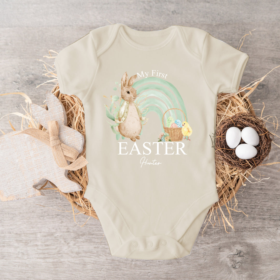 Personalised beige Easter baby vest that says 'My first Easter Hunter'. This design features a sage green rainbow with a rabbit wearing a beige jacket on one side an Easter basket filled with eggs