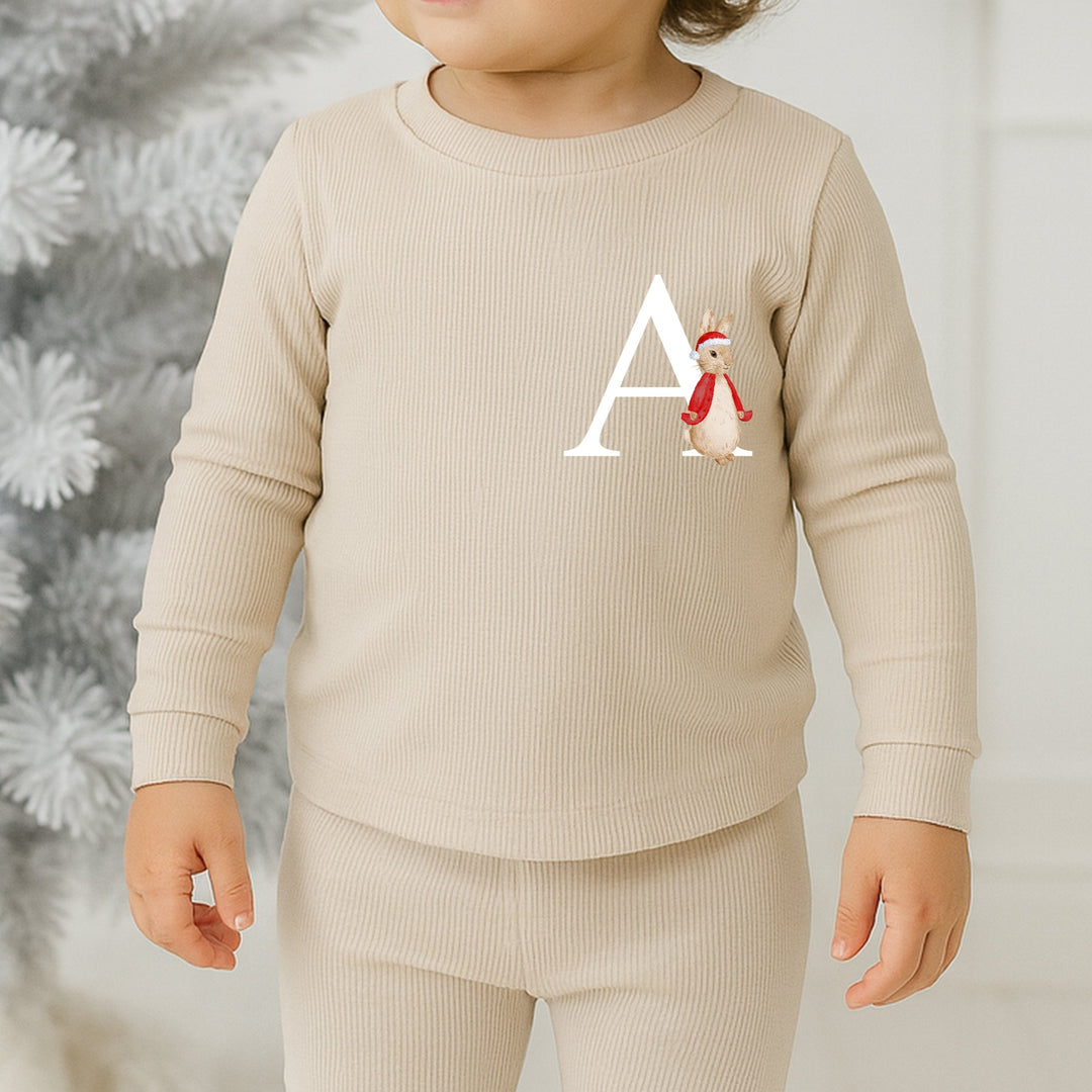 Child wearing a beige outfit with a letter 'A' and a penguin design, standing in front of a decorated Christmas tree.