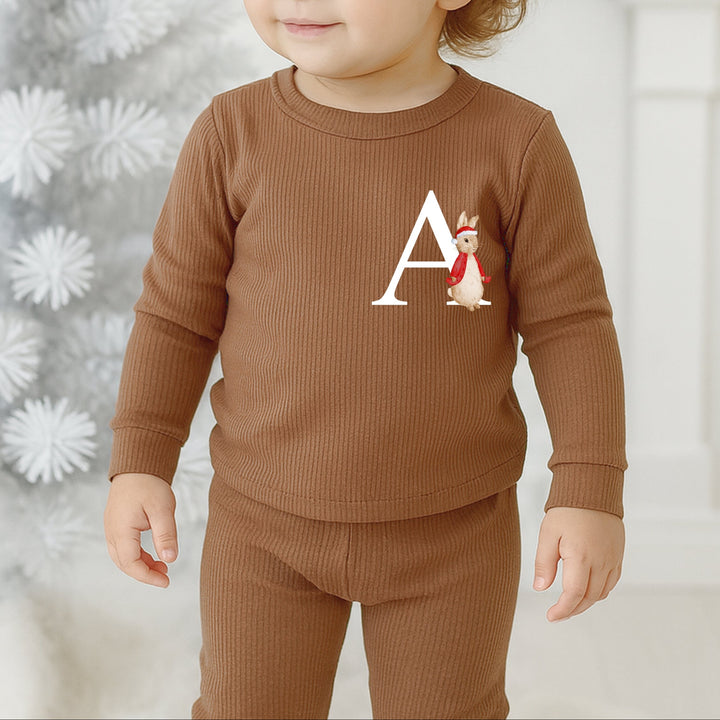 Child wearing a brown outfit with a letter 'A' and cartoon character, standing in front of a decorated Christmas tree.