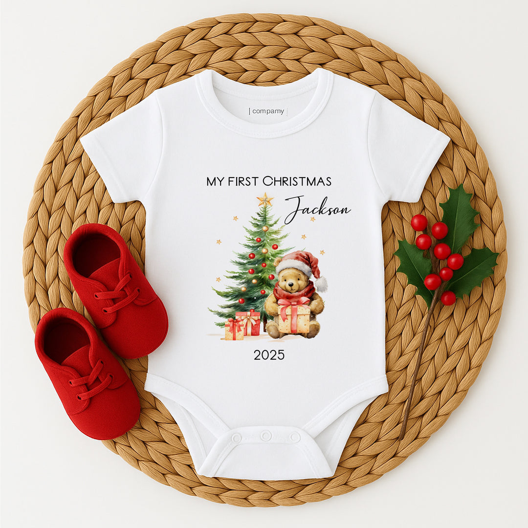Baby onesie with Christmas design and text, red shoes, and holly leaves on a woven mat.