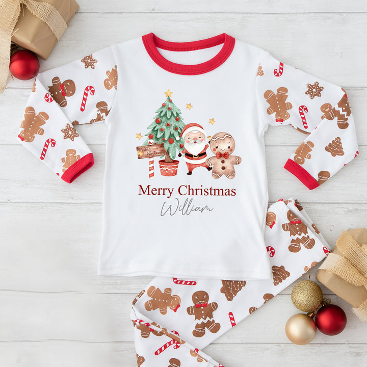 A white long-sleeve Christmas-themed pajama top with a gingerbread man and a Christmas tree, along with personalized text 'Merry Christmas William'. The pajama bottoms are red with festive patterns.