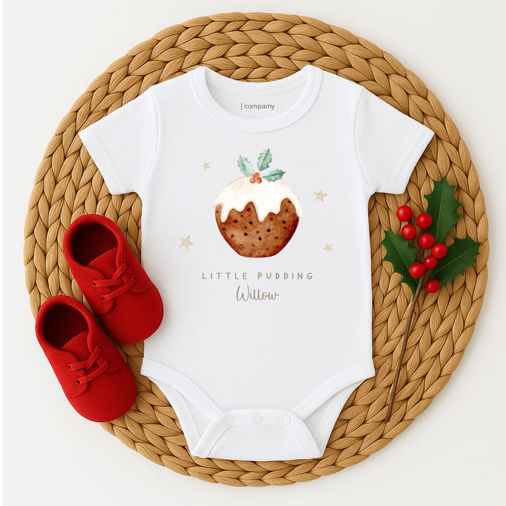 White baby onesie with Christmas pudding design on a woven mat with red shoes and holly.