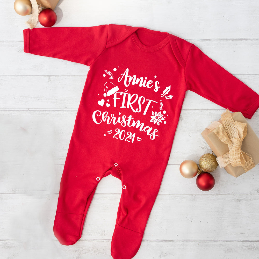 A red bodysuit for babies, with 'Annie's First Christmas 2024' printed on the front, presented on a white wooden background with gold and red decorations.