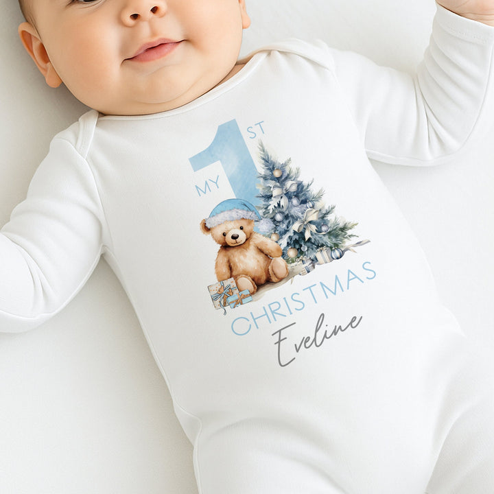 A white baby grow that features a big blue 1 and a teddy bear wearing a blue christmas hat sitting next to a christmas tree. 