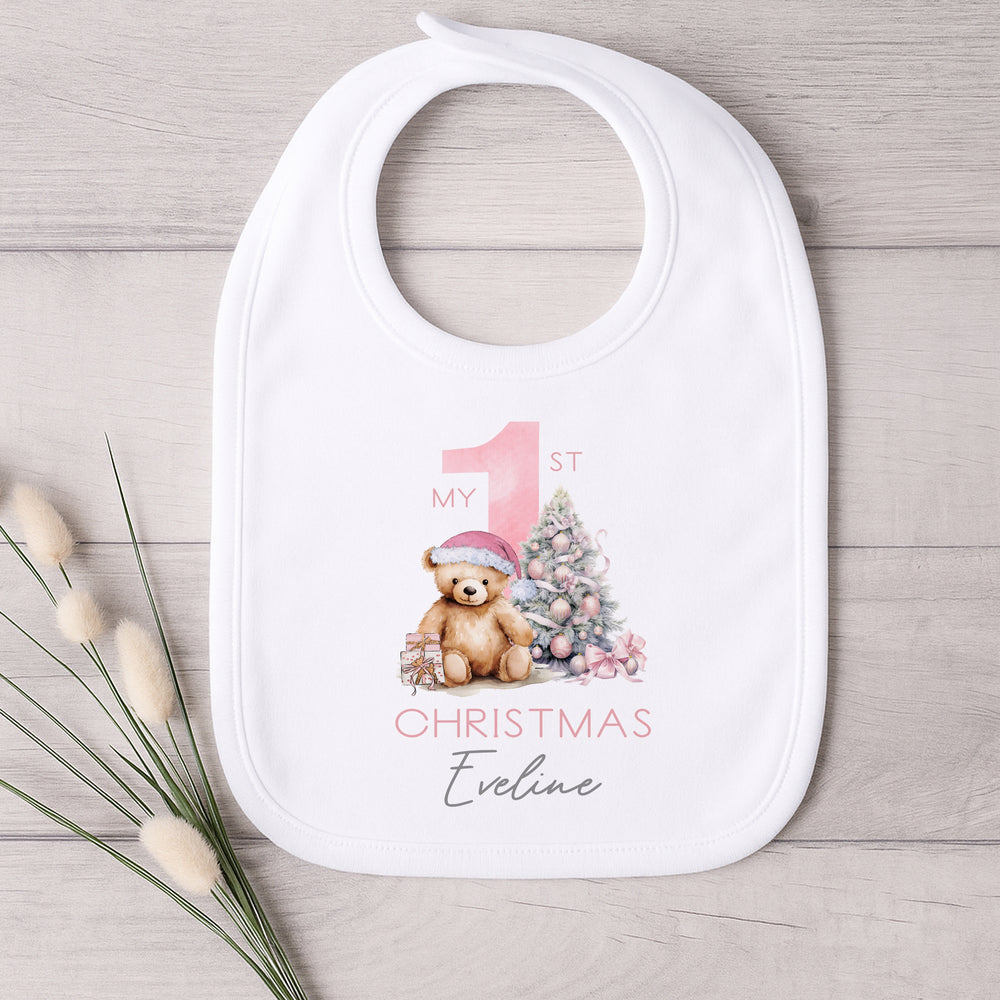 A white baby bib that features a big pink 1 and a teddy bear wearing a pink christmas hat sitting next to a christmas tree. 