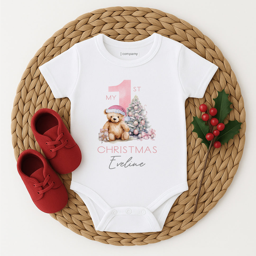 A white baby vest that features a big pink 1 and a teddy bear wearing a pink christmas hat sitting next to a christmas tree. 