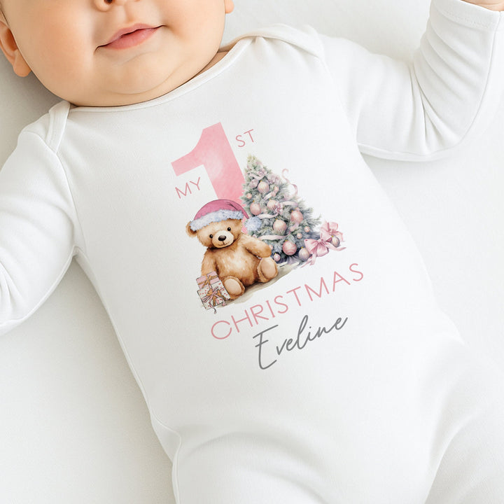 A white baby grow that features a big pink 1 and a teddy bear wearing a pink christmas hat sitting next to a christmas tree. 