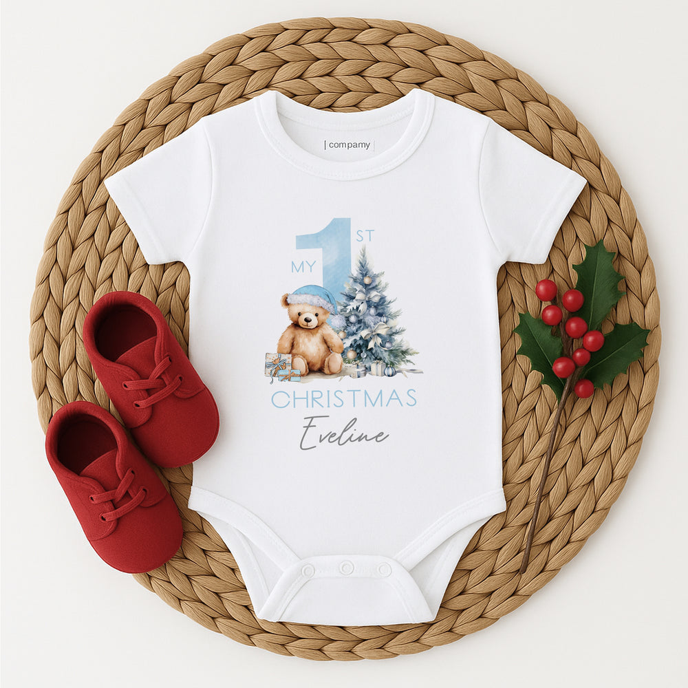 A white baby vest that features a big blue 1 and a teddy bear wearing a blue christmas hat sitting next to a christmas tree. 