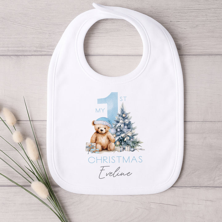 A white baby bib that features a big blue 1 and a teddy bear wearing a blue christmas hat sitting next to a christmas tree. 