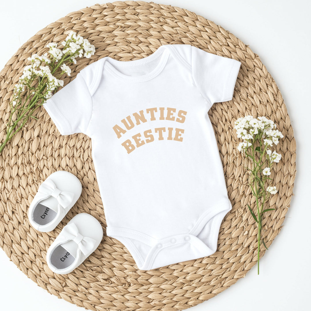 Baby onesie with 'Aunties Bestie' text on a woven mat with baby shoes and flowers.