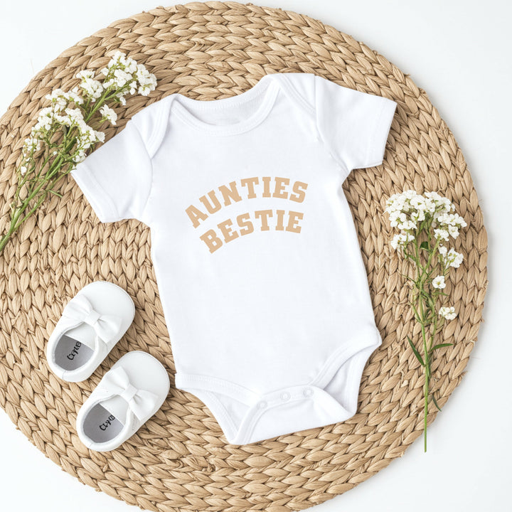 Baby onesie with 'Aunties Bestie' text on a woven mat with baby shoes and flowers.