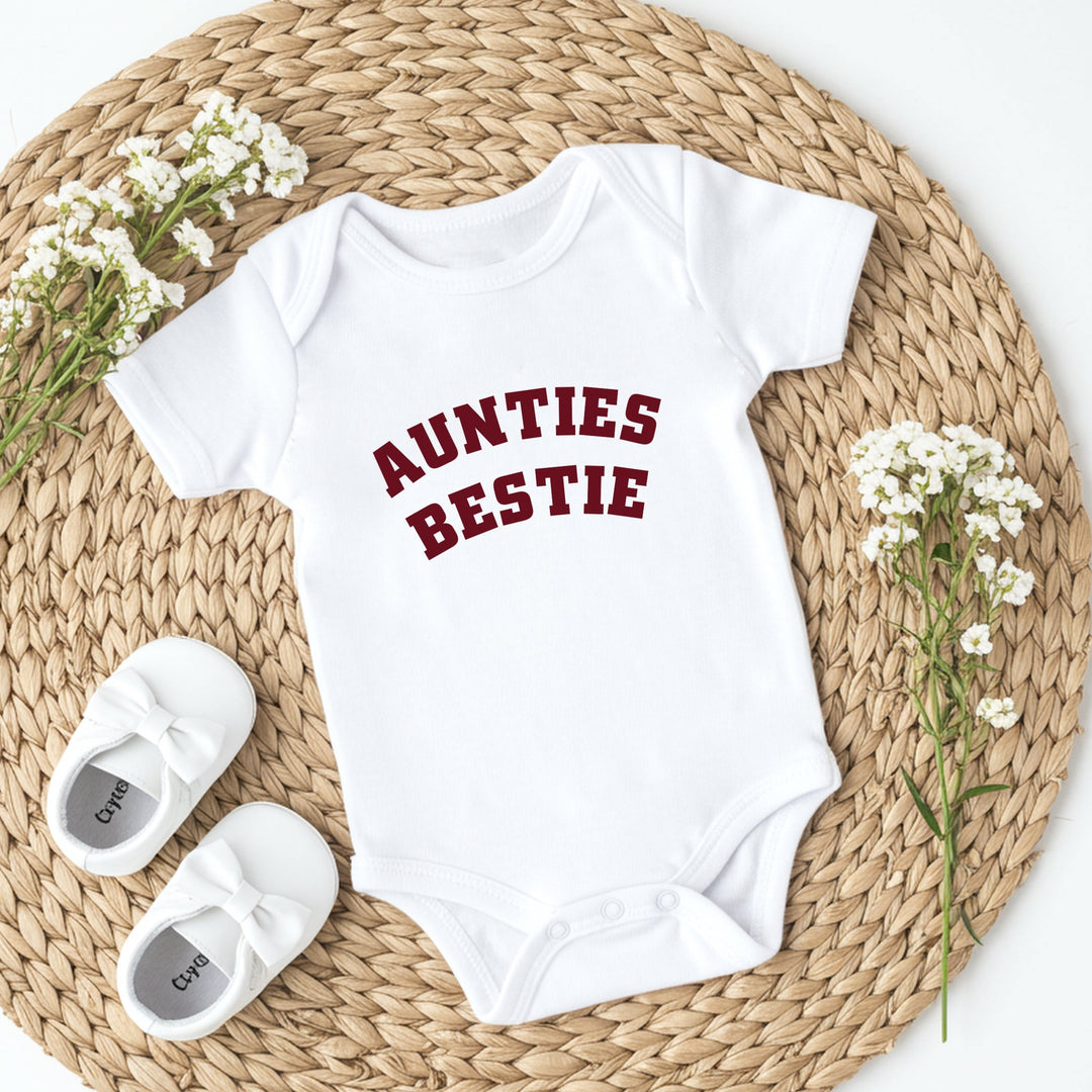 White baby onesie with 'Aunties Bestie' text, white baby shoes, and baby flowers on a woven mat.