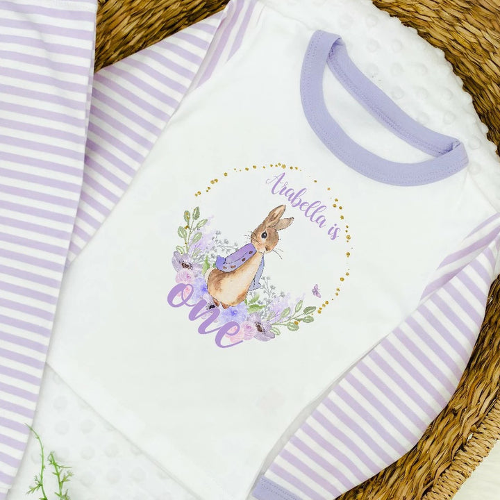 A pair of lilac and white stripe children's birthday pyjamas with a purple floral bunny wreath design saying "Arabella is One".