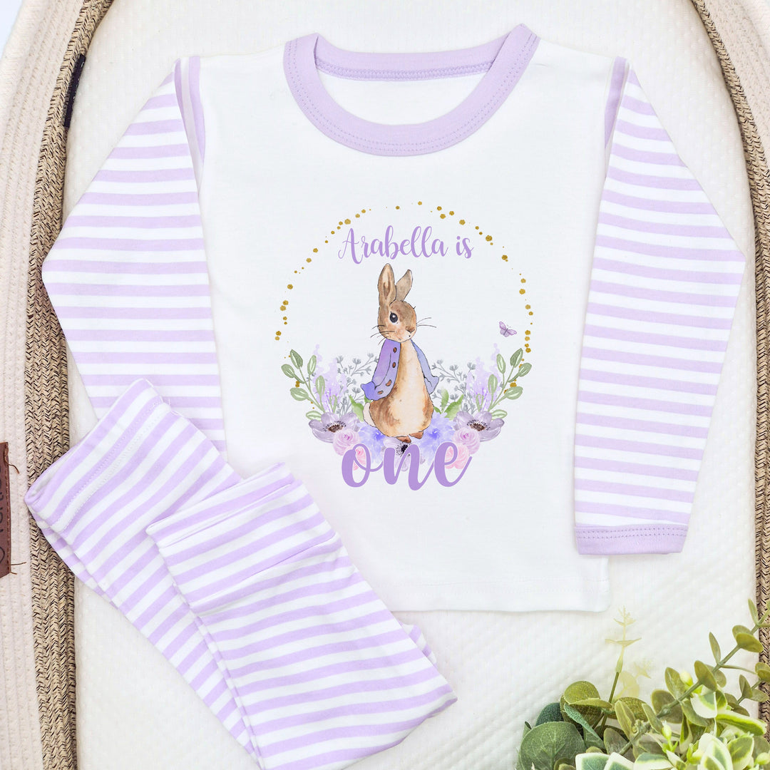 A pair of lilac and white stripe children's birthday pyjamas with a purple floral bunny wreath design saying "Arabella is One".