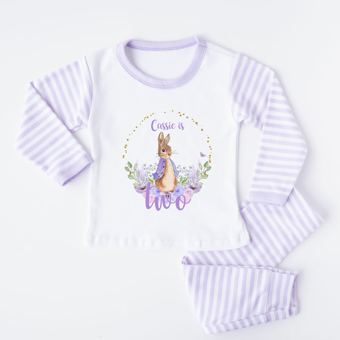 A pair of lilac and white stripe children's birthday pyjamas with a purple floral bunny wreath design saying "Cassie is One".