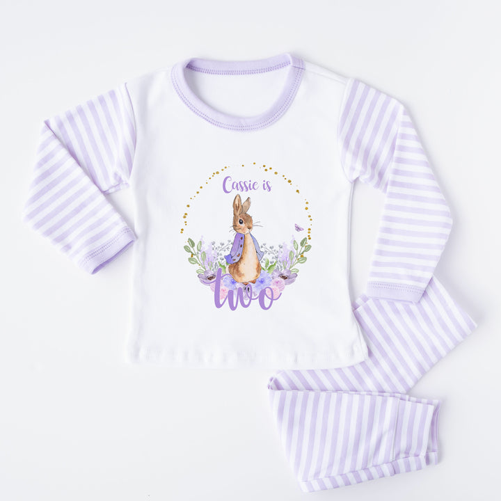 A pair of lilac and white stripe children's birthday pyjamas with a purple floral bunny wreath design saying "Cassie is One".