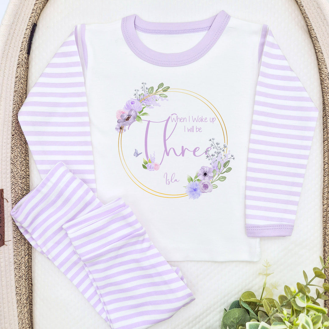 A pair of lilac and white stripe children's birthday pyjamas with a purple floral butterfly wreath design saying "When I wake up I will be One" with the name "Isla".