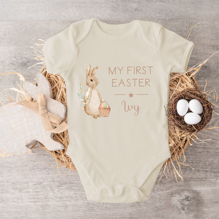 Personalised beige Easter baby vest that says 'My First Easter Oscar'. This design features a rabbit wearing a beige jacket holding an Easter basket full of eggs