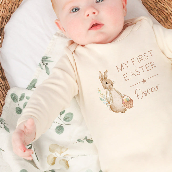 Personalised beige Easter baby grow/sleepsuit that says 'My First Easter Oscar'. This design features a rabbit wearing a beige jacket holding an Easter basket full of eggs