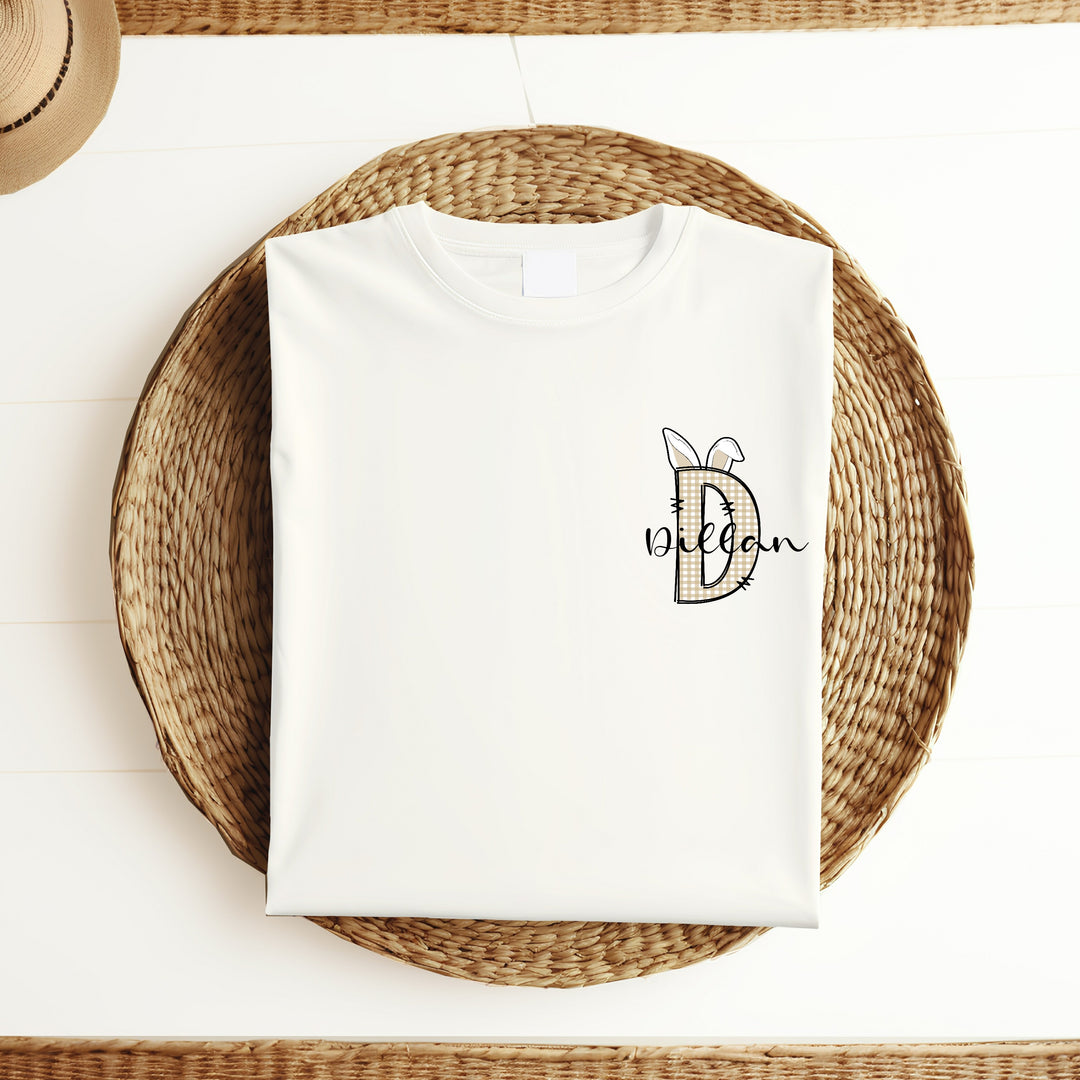 Personalised white Easter t-shirt that says 'D Dillan'. This design features a beige tartan D with bunny ears