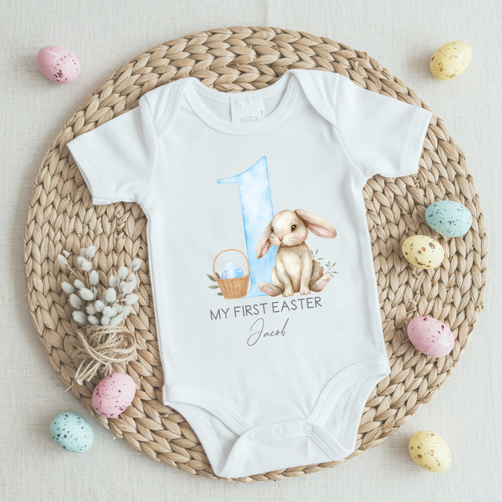 Personalised white baby Easter vest that says 'My First Easter Jacob'. This design features a big blue 1 with a white rabbit oneside and an Easter basket