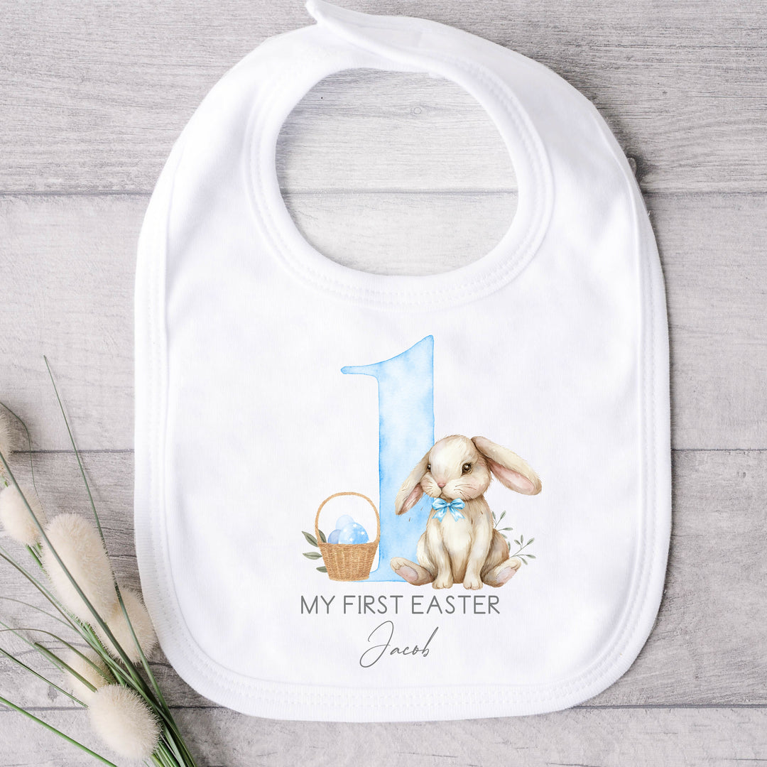 Personalised white baby Easter bib that says 'My First Easter Jacob'. This design features a big blue 1 with a white rabbit oneside and an Easter basket