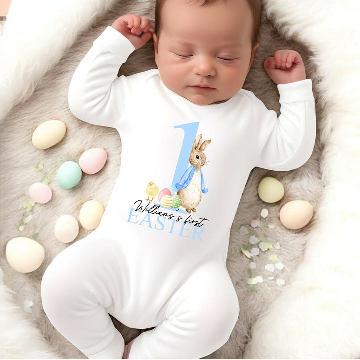 Personalised white Easter baby grow/sleepsuit that says 'William's first Easter'. This design features a big blue 1 and a rabbit wearing a blue jacket standing on one side of the number. On the other side there is Easter eggs and a  yellow chick