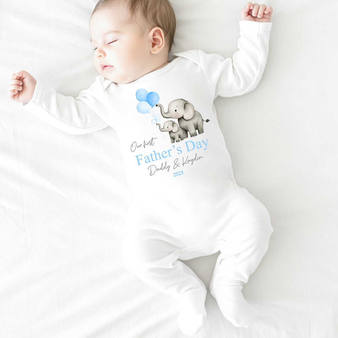 Personalised white Father's Day baby grow/sleepsuit that says 'Our First Father's Day Daddy & Kaydin 2025'. This design features 2 grey elephants holding 3 blue balloons