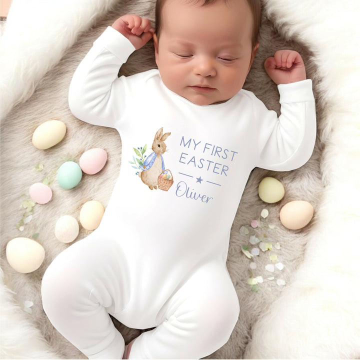 Personalised white Easter baby grow/sleepsuit that says 'My First Easter Oliver'. This design features a rabbit wearing a blue jacket holding an Easter basket full of eggs